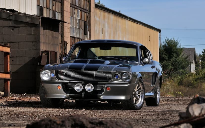 Silver Shelby GT500 Eleanor muscle car with black racing stripes parked in front of a rustic industrial building.