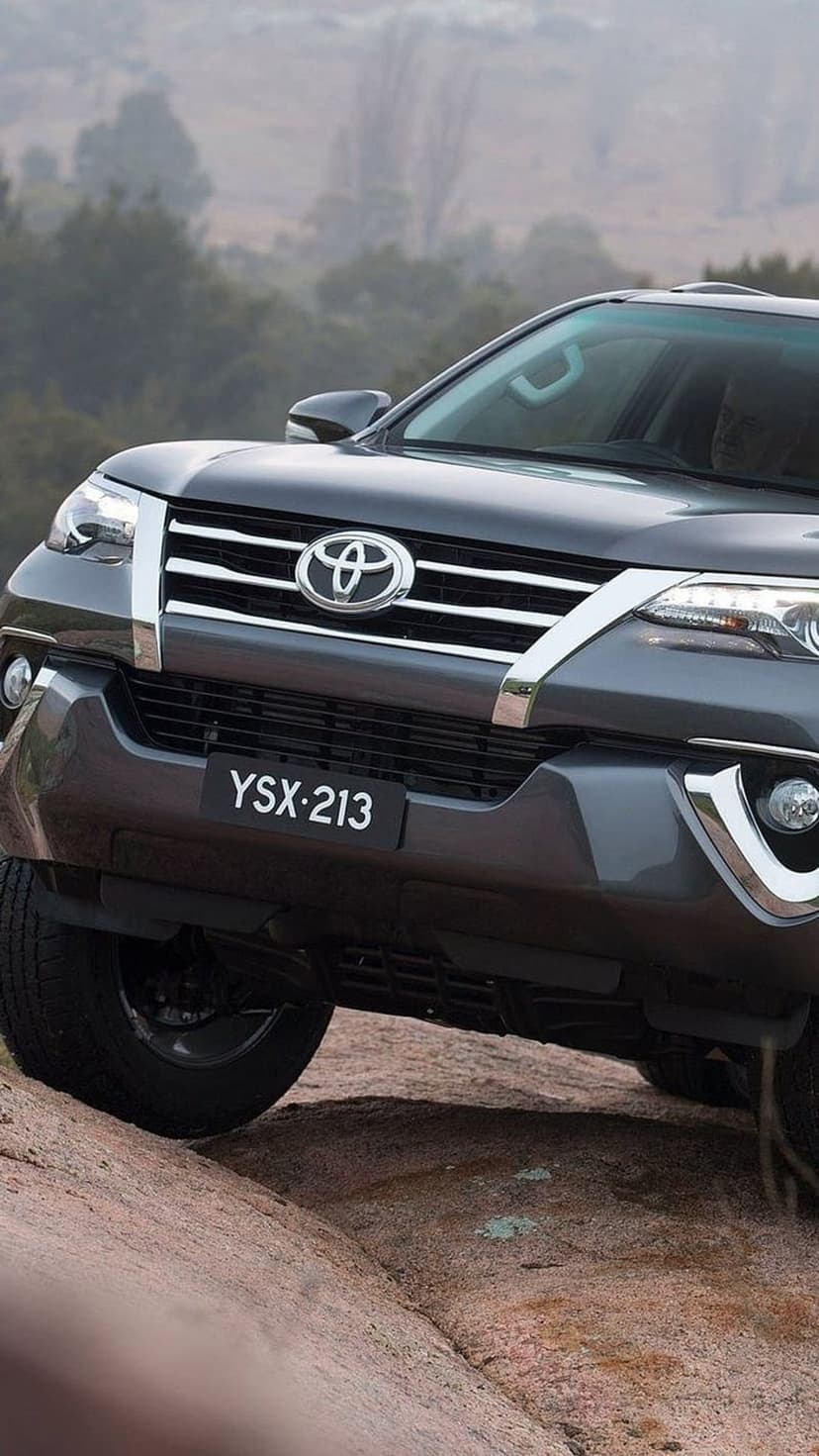 Toyota Fortuner SUV with headlights on navigates a steep rocky trail in a misty, pine-covered forest area.