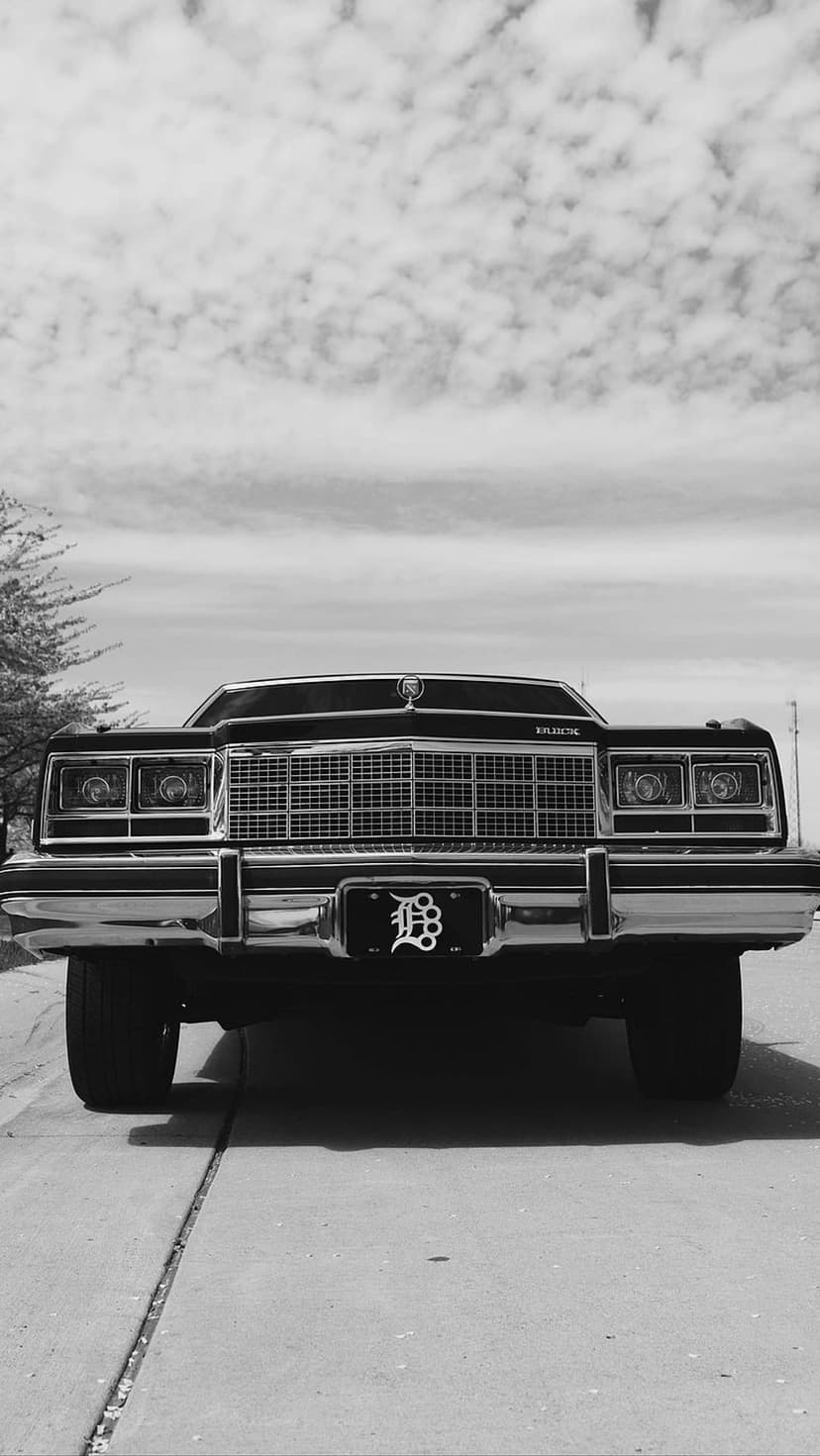 Chrome Buick grille and dual headlights from a low angle against a cloudy sky in a vertical monochrome photo.