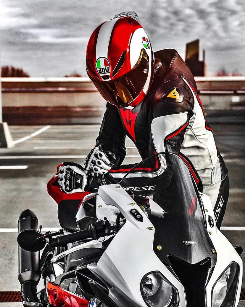 Motorcyclist in an AGV helmet and Dainese suit leans on a white sportbike against a cloudy sky on asphalt.