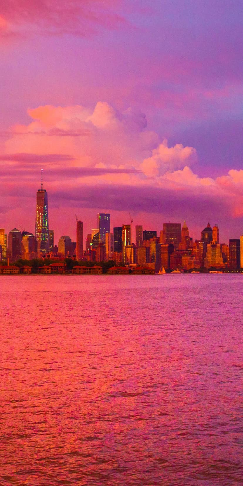 Manhattan skyline silhouettes against a vibrant pink and purple sunset with reflections on the calm water surface.