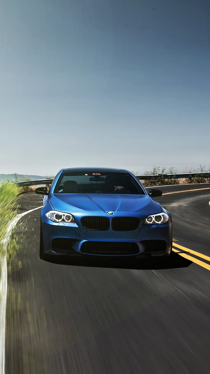 Blue BMW M5 sedan drives on a winding asphalt road surrounded by green trees under a clear sky during the daytime.