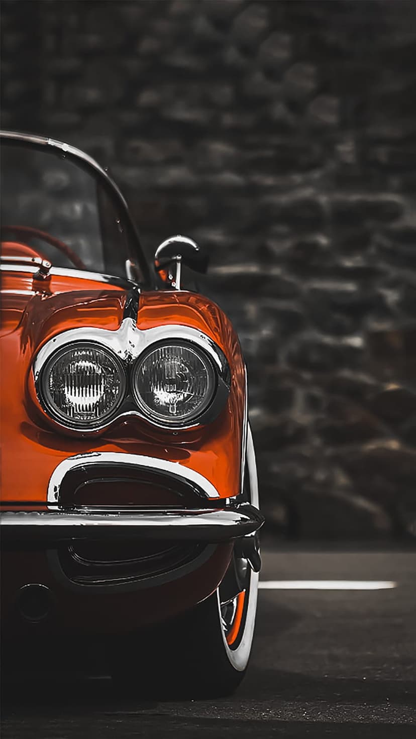 Orange Corvette front end features dual headlights and reflective chrome bumpers against a dark stone wall.