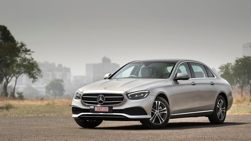 Silver Mercedes-Benz E-Class sedan parked on a gravel lot with a hazy, blurred city skyline background.