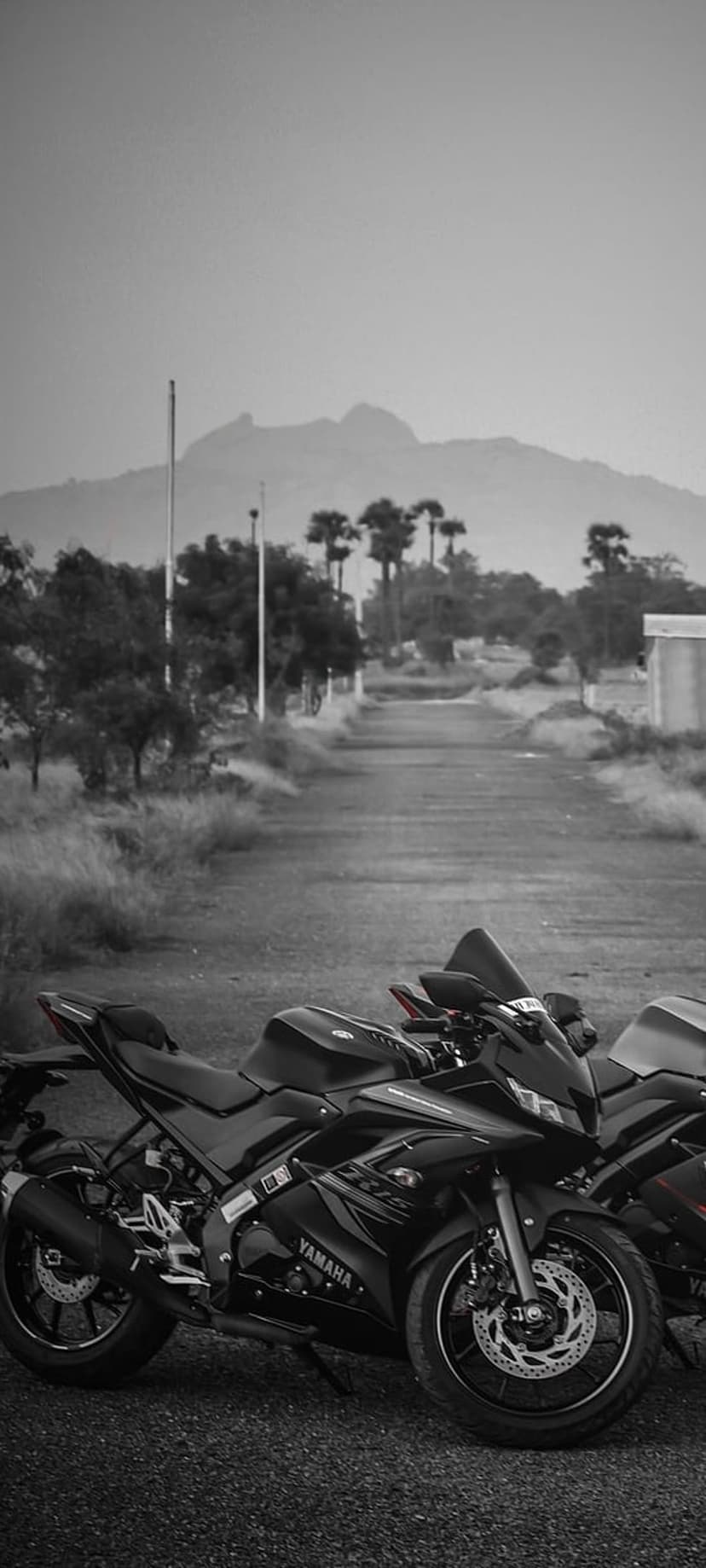 Two black Yamaha R15 motorcycles parked on an asphalt road with mountains and palms in a monochrome landscape.