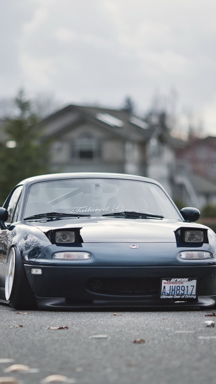 Front view of a stanced dark blue Mazda Miata with pop-up headlights and white wheels on a residential street.