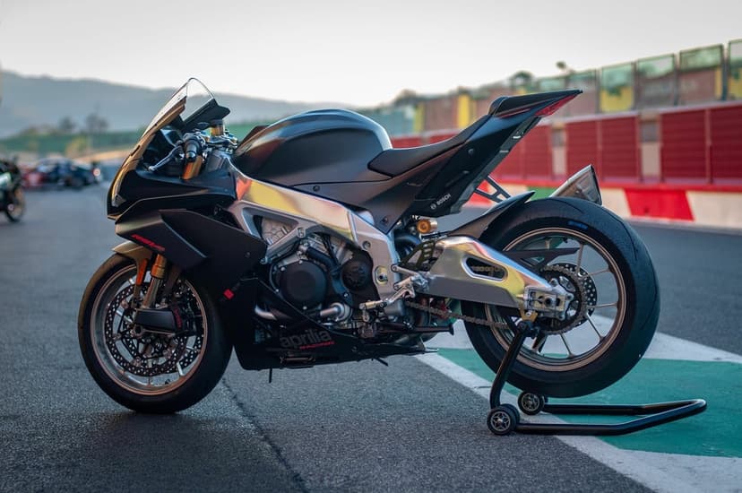 Matte black Aprilia RSV4 motorcycle on a paddock stand at a racetrack during sunset with soft bokeh hills.