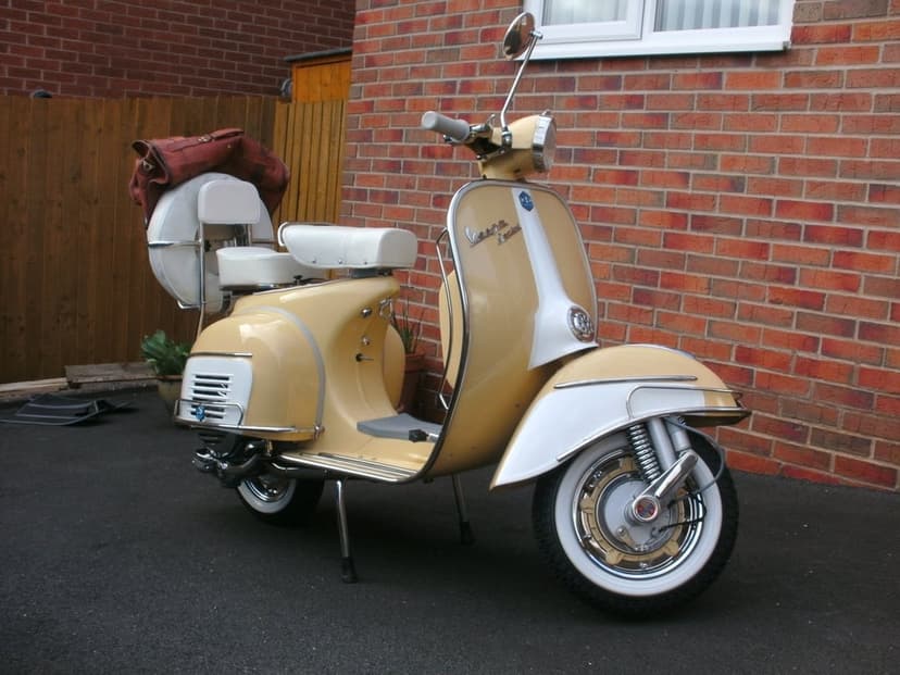 Vintage cream and white Vespa scooter with chrome trim and whitewall tires parked against a red brick wall.