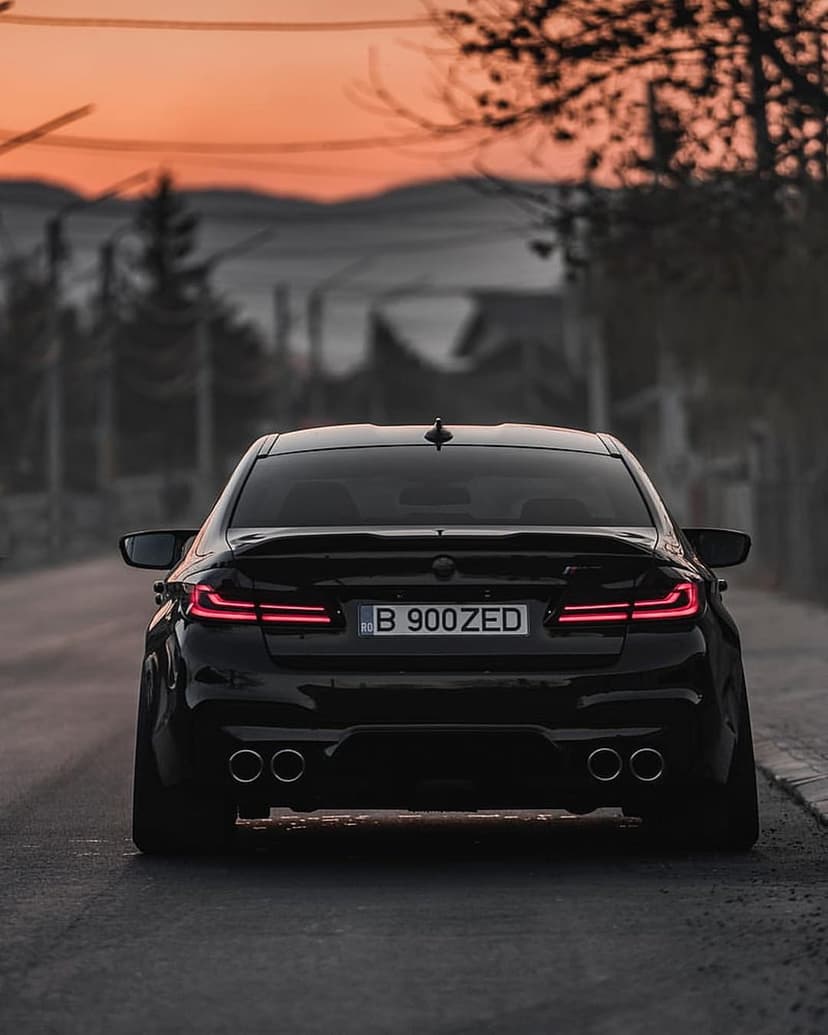 Black BMW M5 rear view with glowing red taillights on a road under a vibrant orange and purple sunset sky.