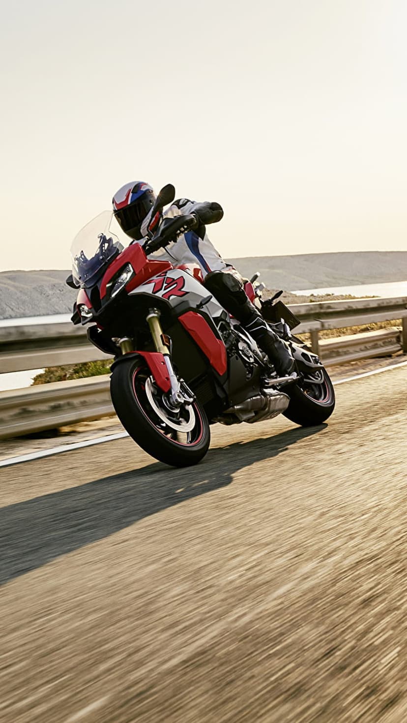 Red sport-touring motorcycle leans into a sharp asphalt turn with a calm blue lake and hazy shore in the background.