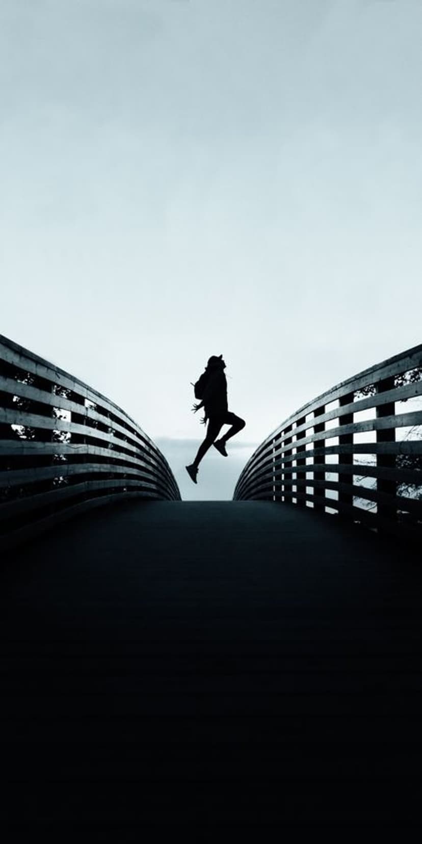 Athletic person in silhouette leaps mid-air between dark bridge railings against a bright minimalist sky.