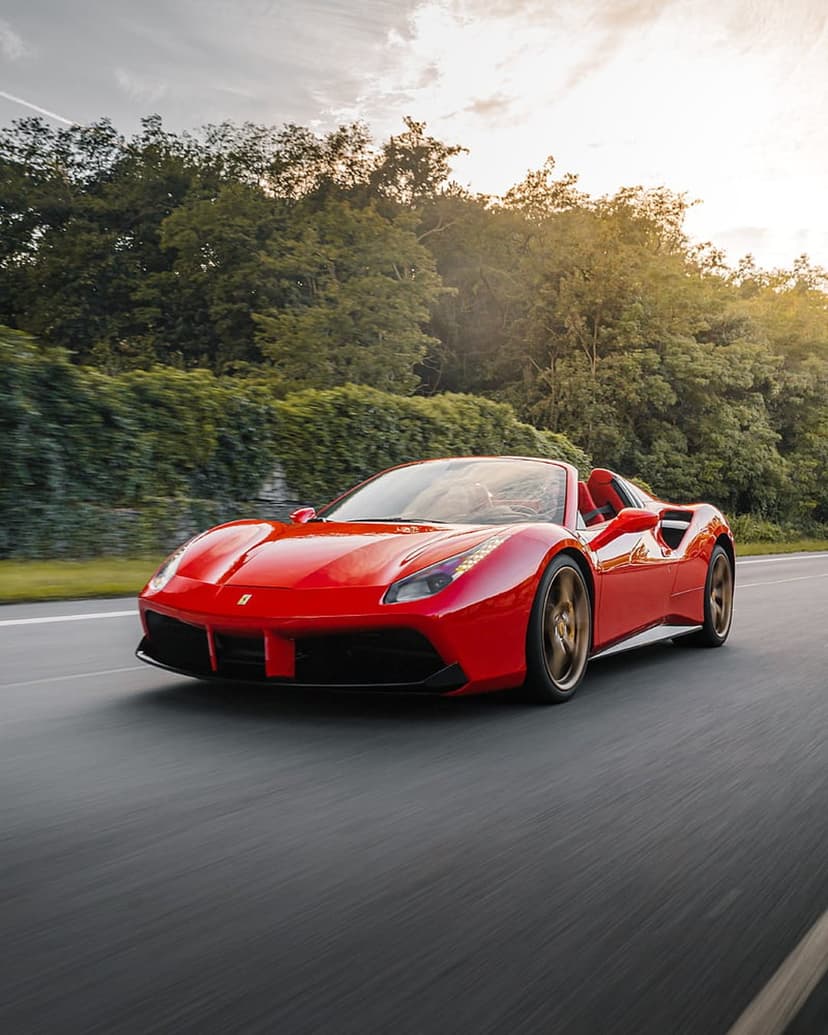 Red Ferrari convertible driving on a tree-lined road at sunset with motion blur and golden hour lighting.