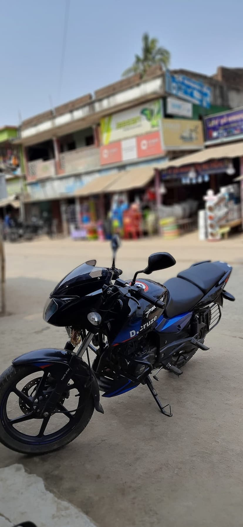Black Bajaj Pulsar D.cruze motorcycle with blue accents parked on a paved street in front of colorful shops.