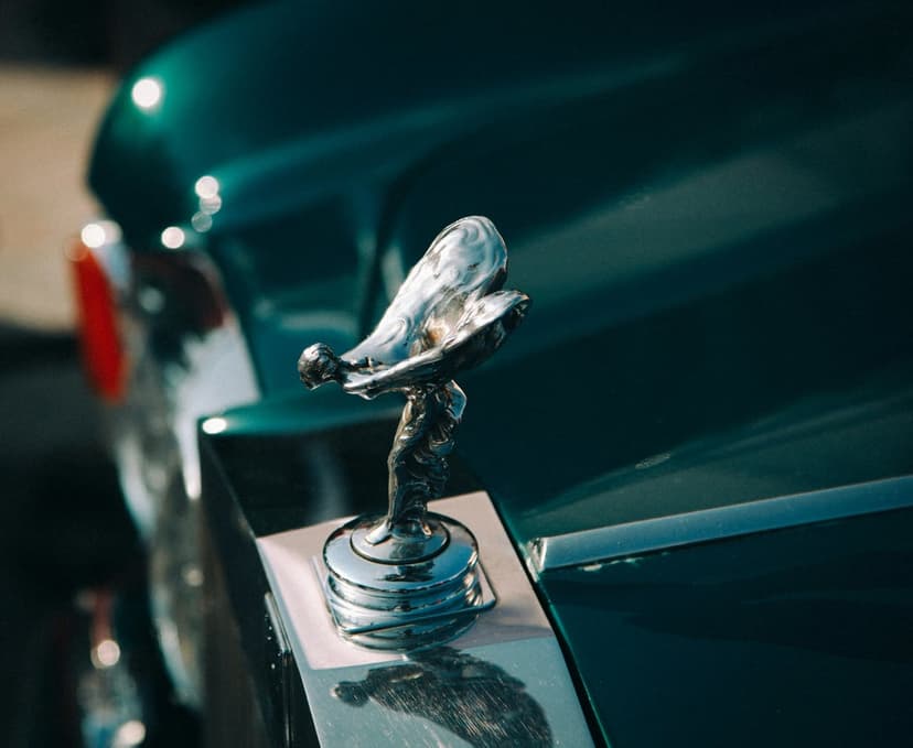 Spirit of Ecstasy figurine in polished chrome stands on a dark green car hood with soft sunlight reflections.