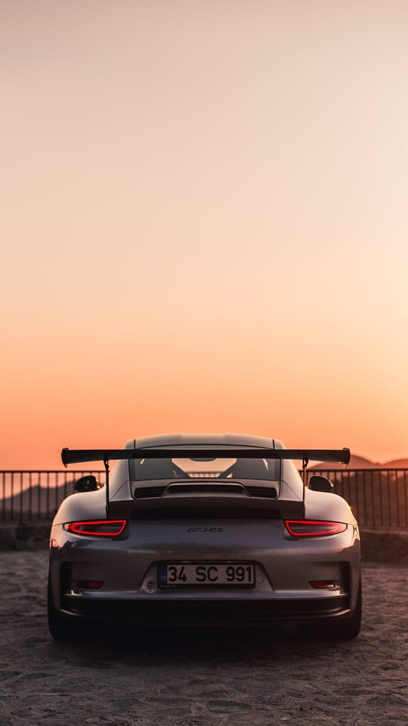 Porsche GT3 RS rear view showing a large wing and red taillights against a sunset sky in vertical orientation.