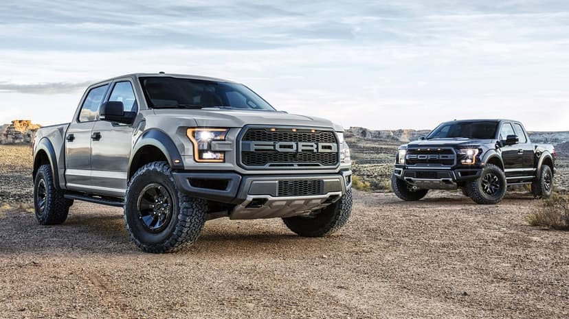 Grey and black Ford F-150 Raptor trucks parked on a dusty desert trail under a cloudy sky with rocky hills.