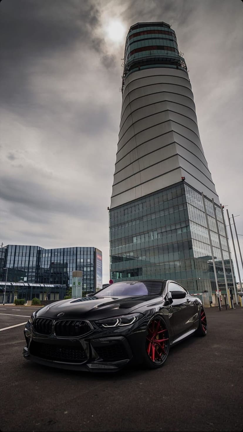 Black BMW M8 coupe with red wheels parked before a curved glass skyscraper under a moody, overcast urban sky.