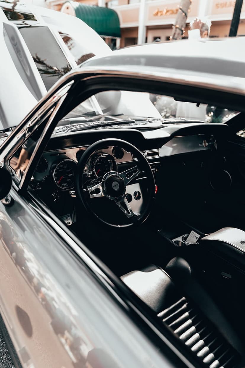 Chrome steering wheel and vintage dashboard gauges inside a silver car with an open hood visible in the background.