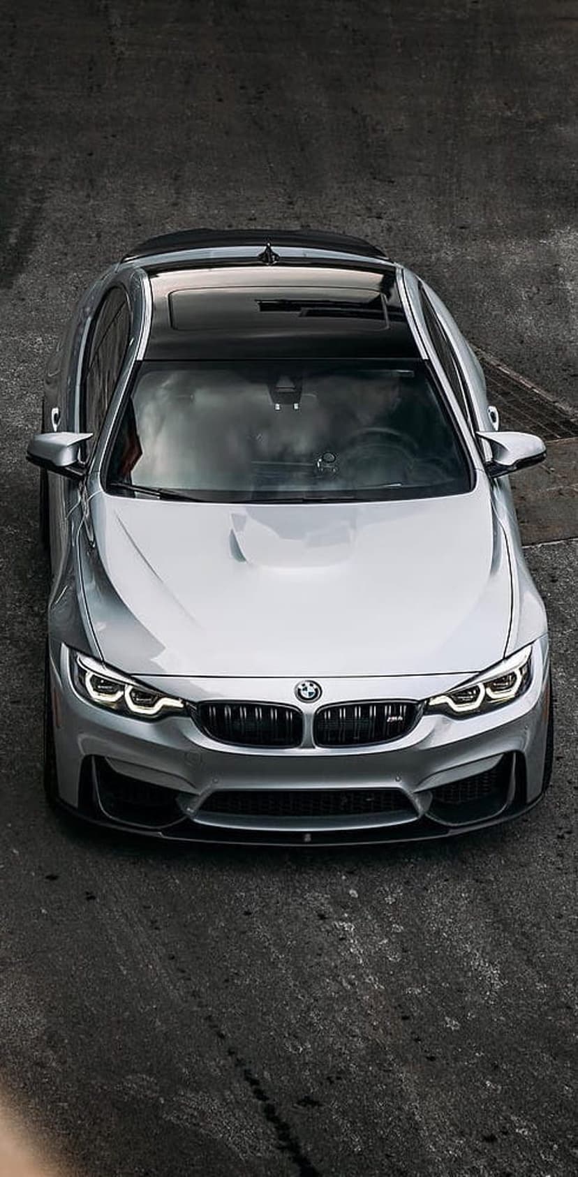 Silver BMW M4 convertible with its top down parked on asphalt, seen from a high-angle aerial perspective.
