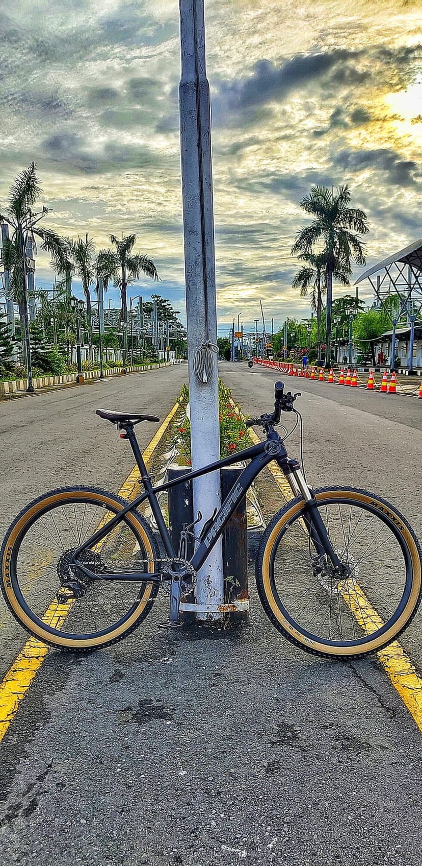 Mountain bike with tan tires leans on a utility pole along a tropical road under a dramatic, cloudy sunset sky.