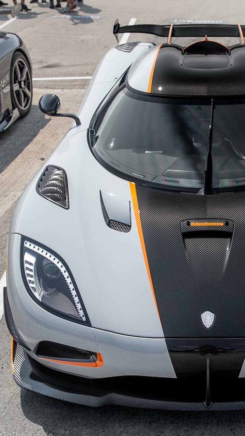 Koenigsegg hypercar featuring white paint, carbon fiber hood, orange accents, and shield emblem on asphalt ground.