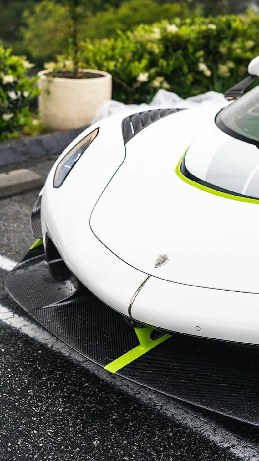 White hypercar front end with lime green accents and carbon fiber splitter against a blurred garden backdrop.