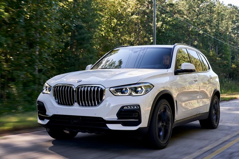 White BMW X5 luxury SUV driving on a tree-lined road with motion blur and natural sunlight.