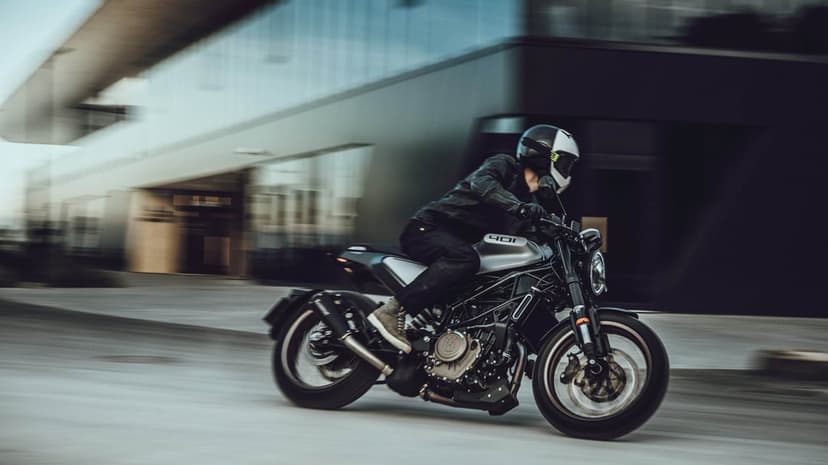 Motorcyclist on a silver Husqvarna 401 riding through an urban area with motion blur and modern architecture.