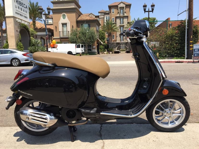 Black Vespa Primavera scooter with tan seat parked on a sidewalk in front of luxury apartment buildings.