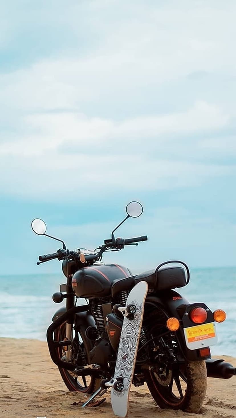 Black motorcycle with red accents and a white skateboard parked on a sandy beach beside the calm blue ocean waves.