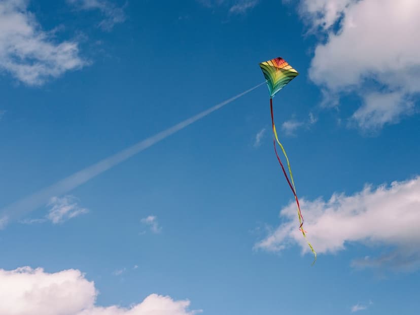Rainbow-colored kite with a long tail soaring past thin white clouds in a bright blue summer sky during the day.