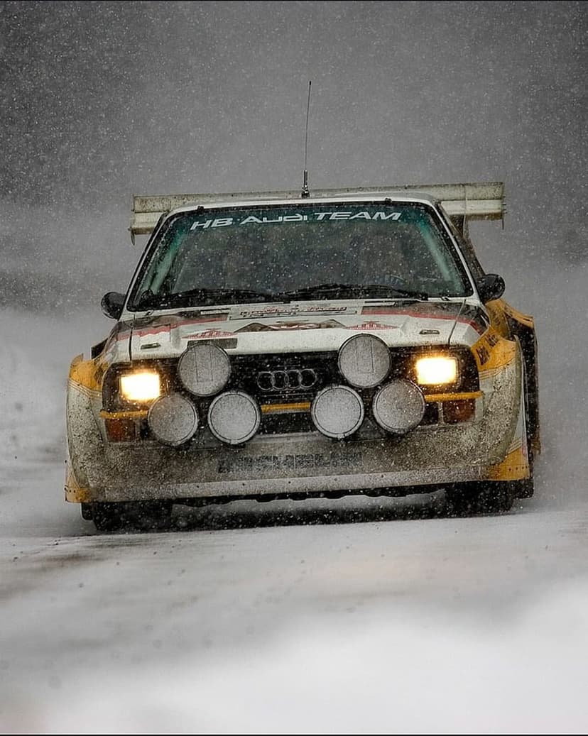 White Audi Quattro rally car with yellow headlights racing through a blizzard on a snowy, mud-splattered track.