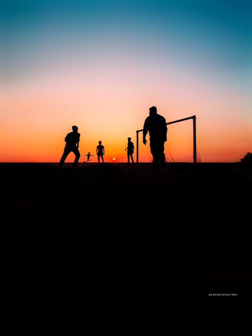 Silhouetted soccer players kick a ball toward a goal against a bright orange and blue sunset sky on a field.
