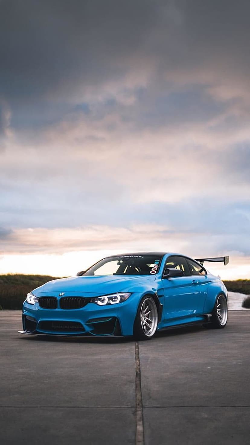 Blue BMW M4 Coupe with a large race spoiler parked on concrete under a cloudy, overcast coastal sky.