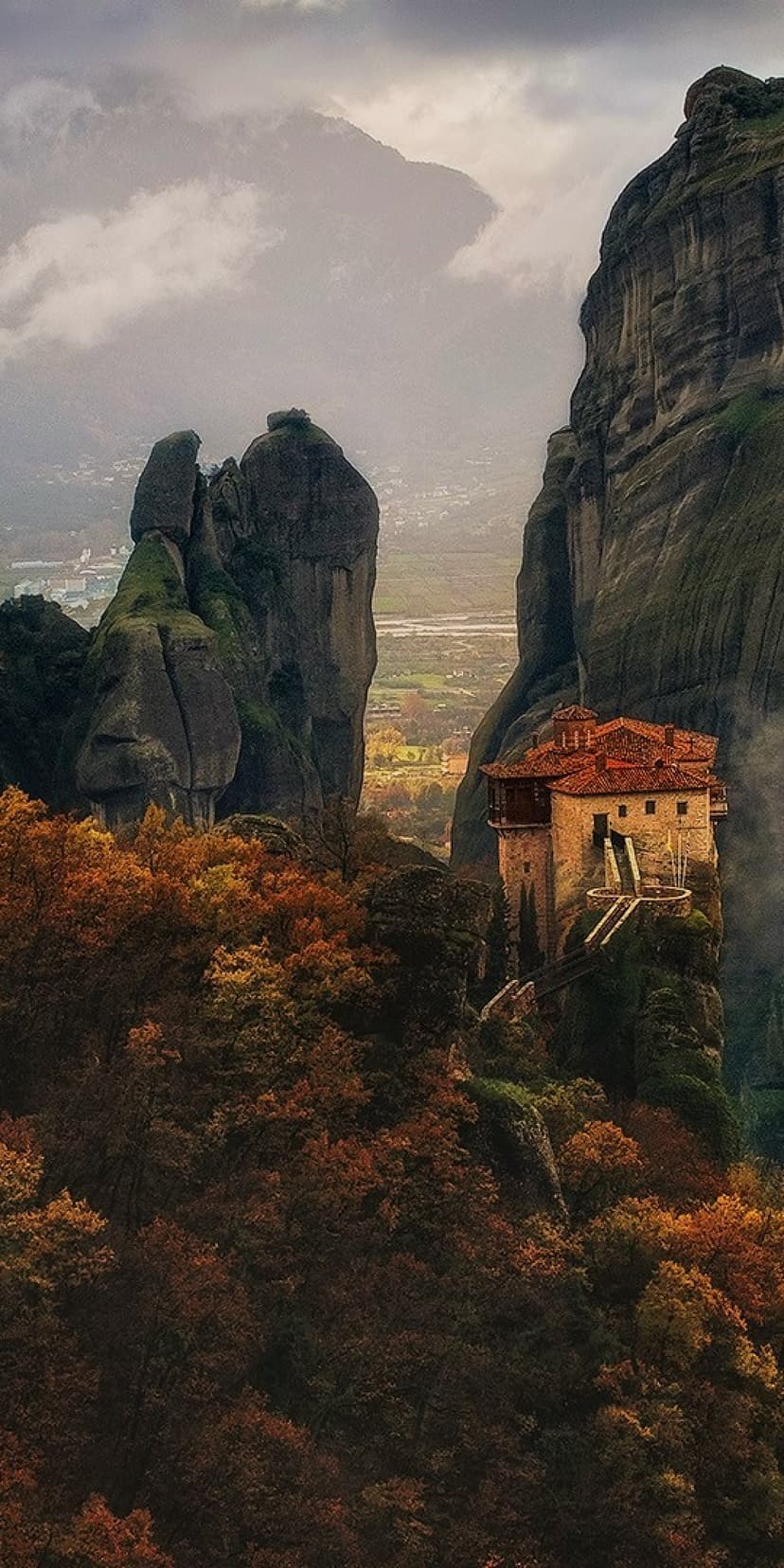 Red-roofed monastery perched on a tall rock cliff surrounded by yellow autumn trees and misty grey mountains.