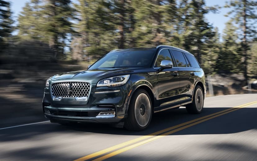 Dark green luxury SUV driving on a paved road with motion-blurred pine trees and bright sunlight.