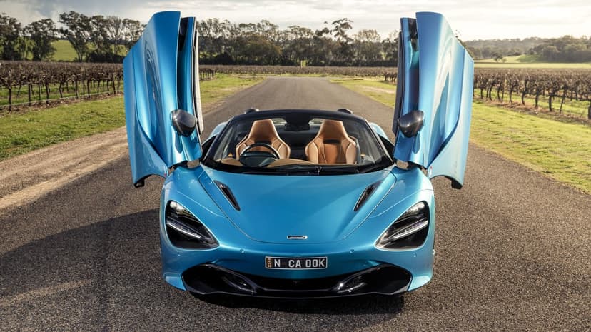 Metallic blue McLaren 720S Spider with open doors parked on a rural road between lush vineyards under a clear sky.