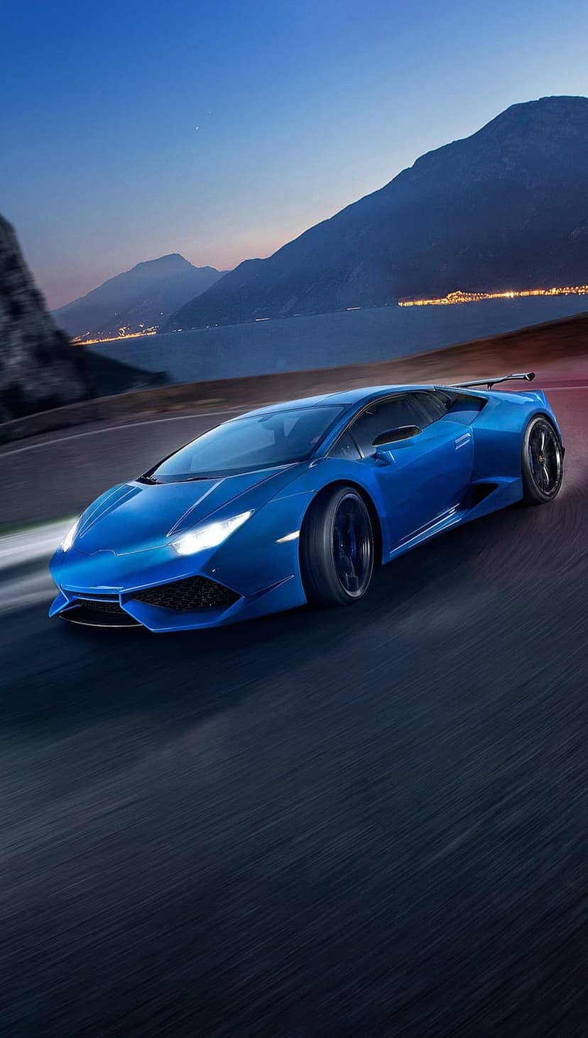 Blue Lamborghini supercar driving on a winding mountain road at dusk with city lights and water in the background.