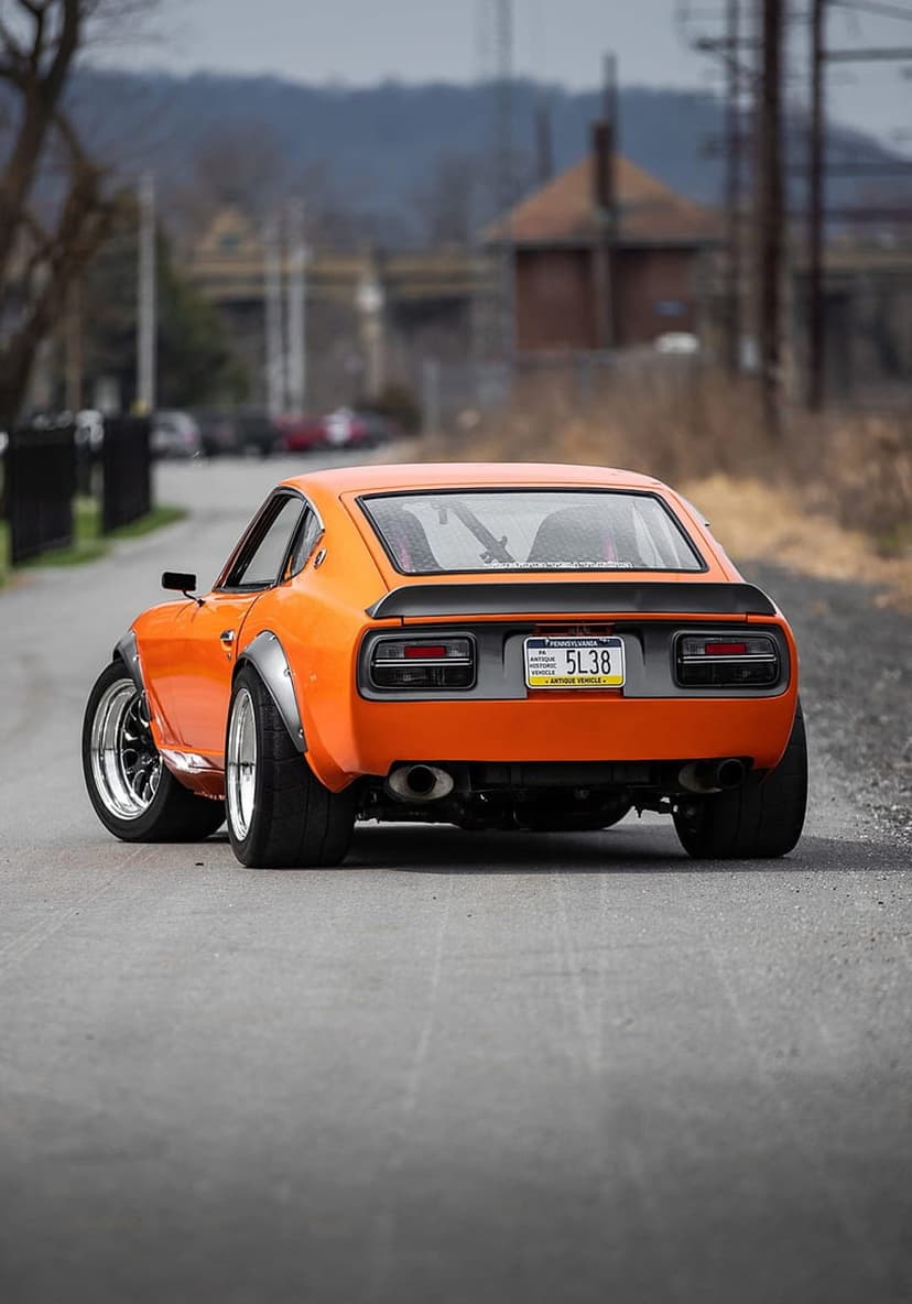 Orange Nissan 240Z with widebody fenders and a rear spoiler parked on a road, shown from a low rear perspective.