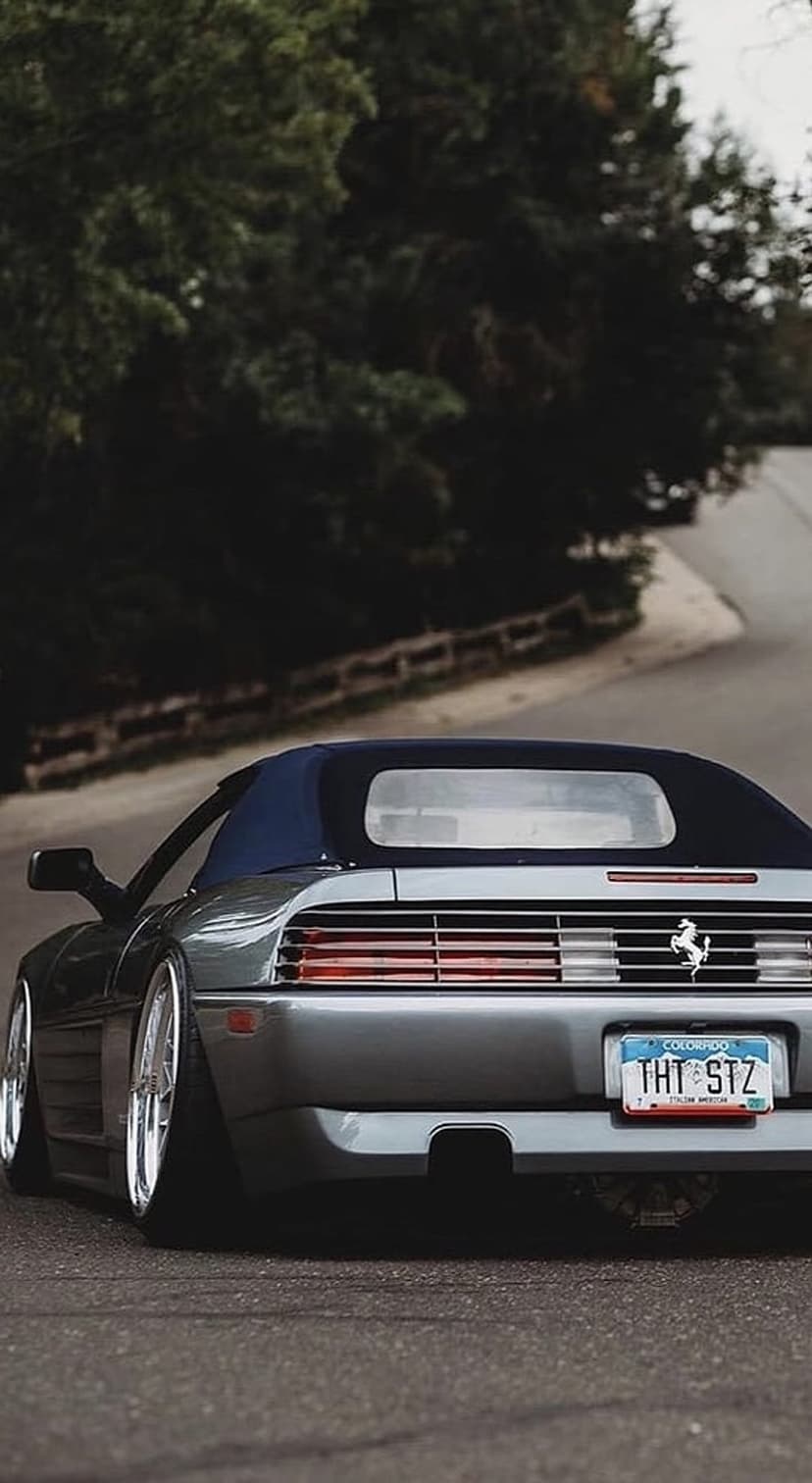Gray Ferrari convertible with a blue top drives on a forest road featuring a custom Colorado license plate.