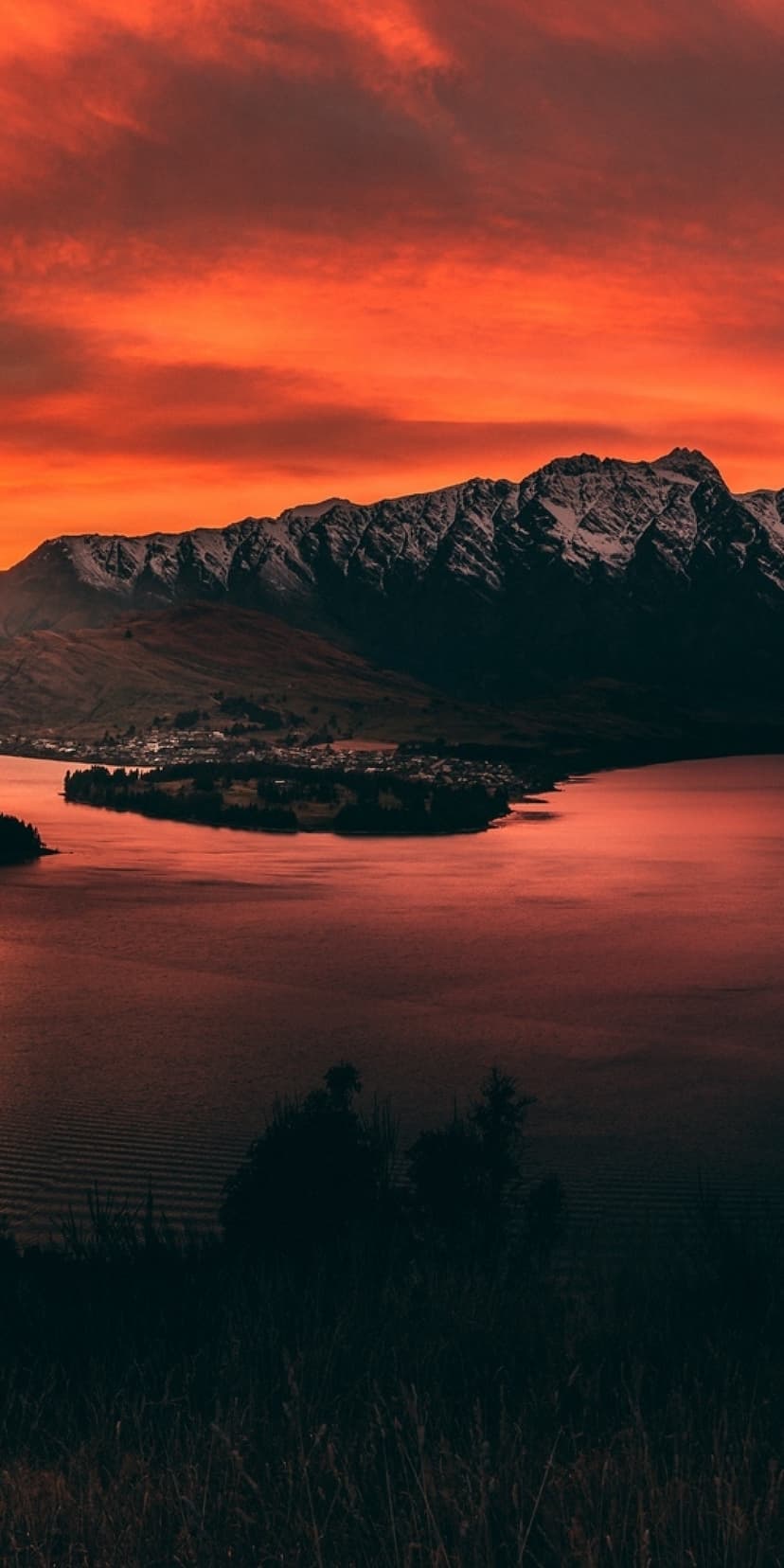 Queenstown buildings nestled beside a lake under a glowing orange sunset and dark mountain silhouettes.