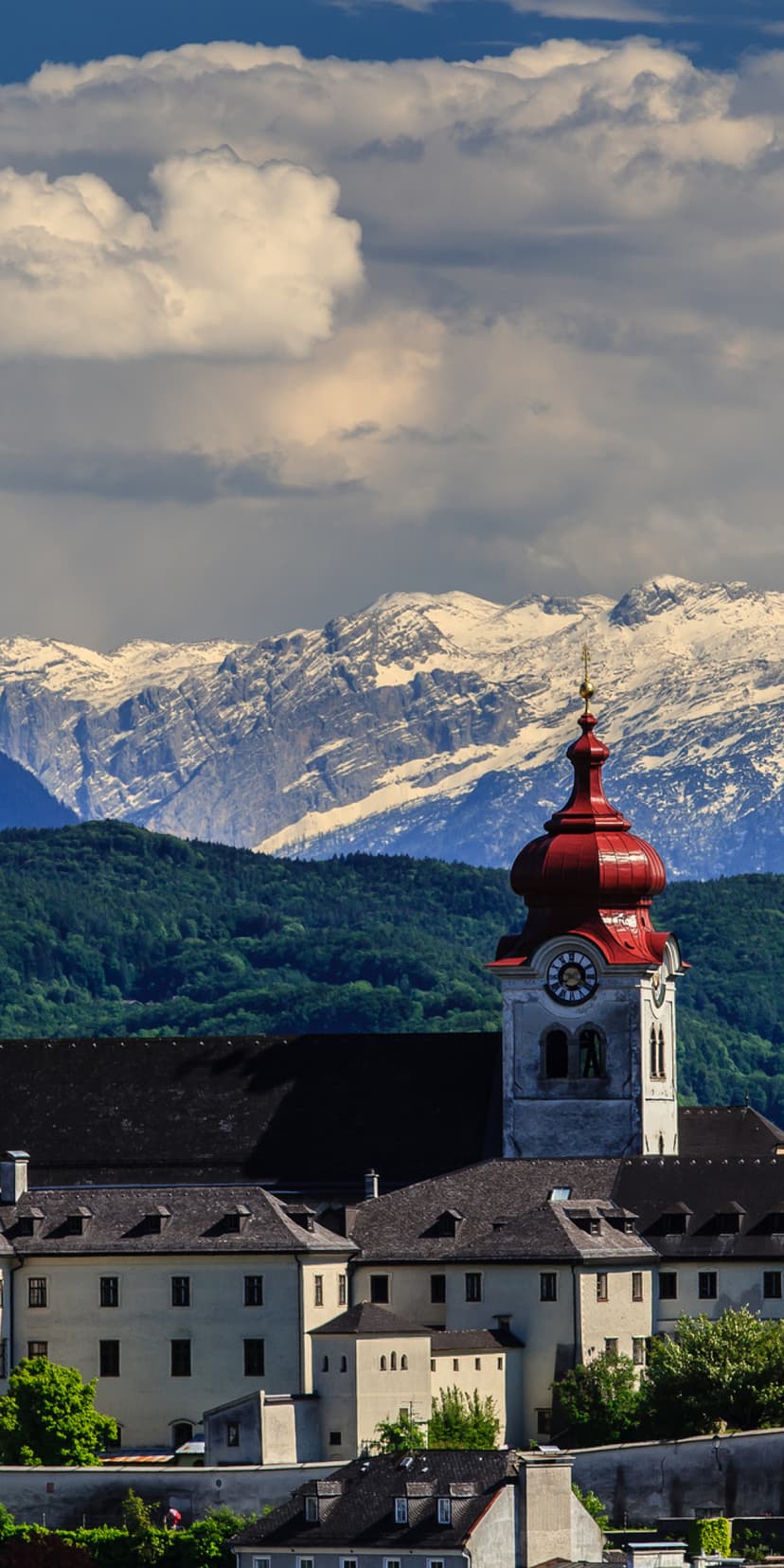 Red-domed steeple of Nonnberg Abbey standing against snow-capped mountains and green trees under a bright blue sky.