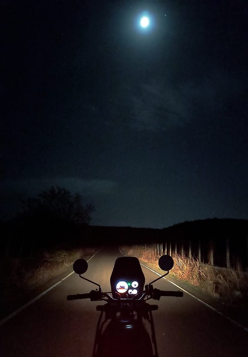 Motorcycle handlebars and glowing dashboard on a dark road under a large bright moon in a cloudy night sky.