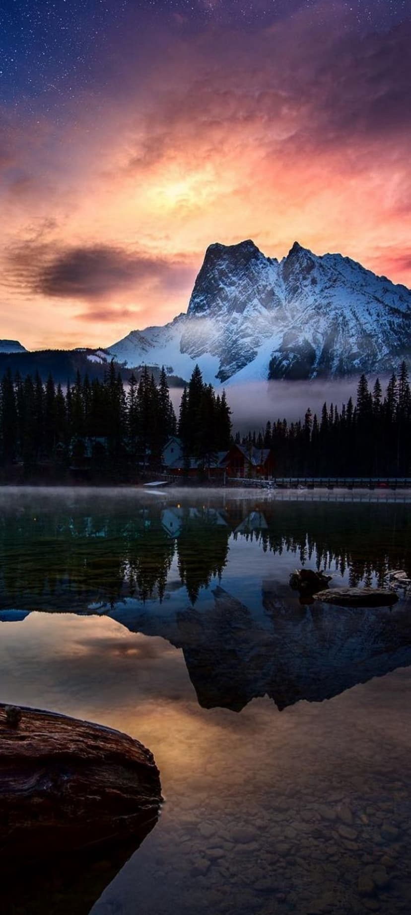 Snow-capped mountains reflect in a still lake under a starry pink sunset with small cabins along the pine forest.