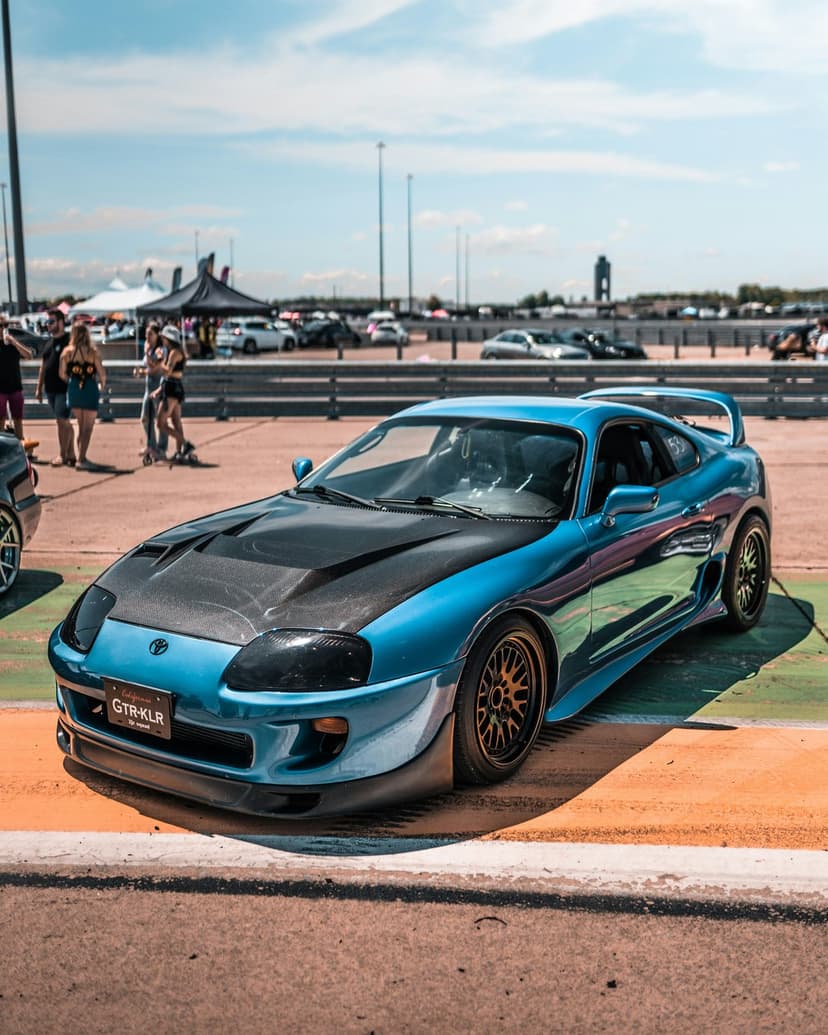 Blue Toyota Supra with a dark carbon fiber hood parked on asphalt with spectators in a blurred background.