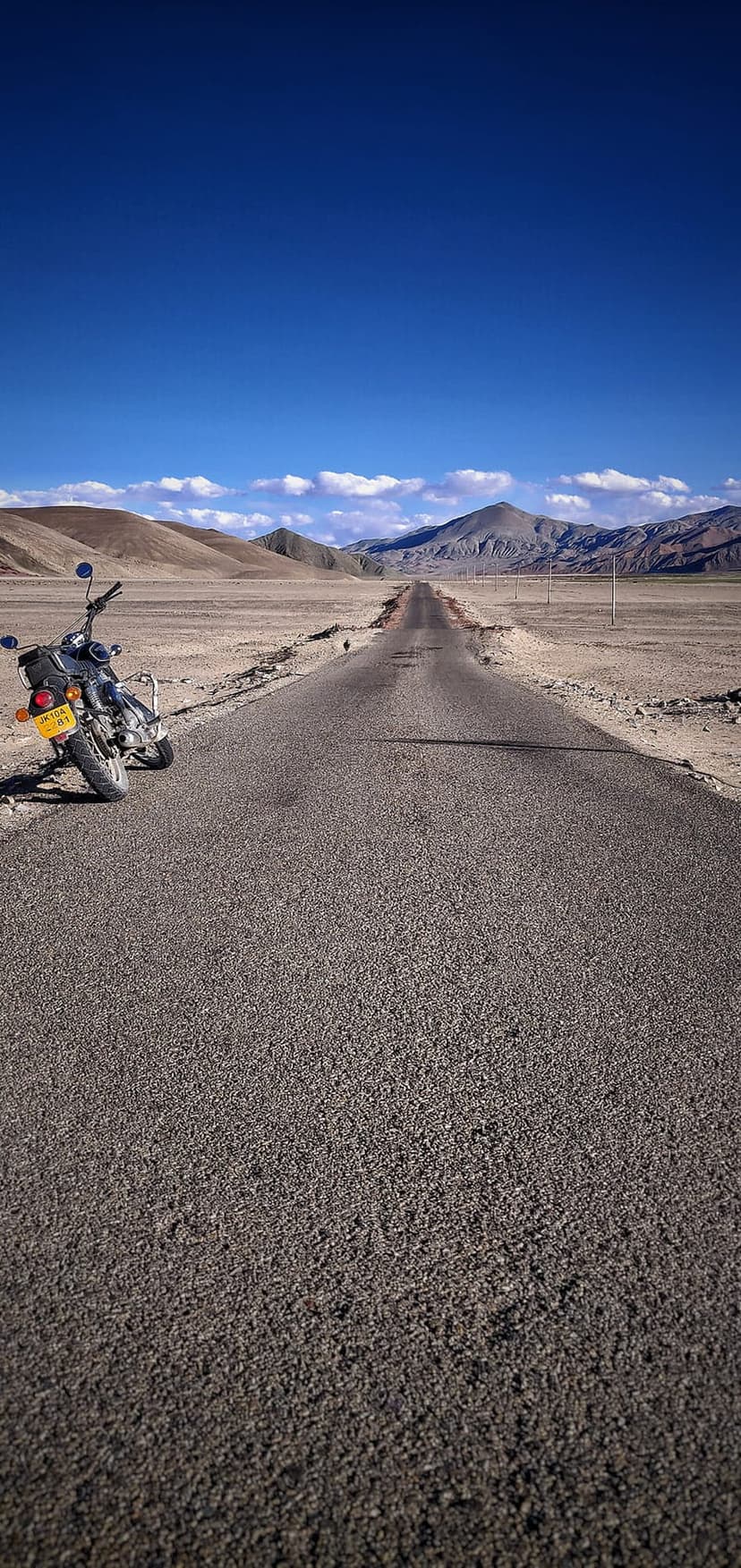 Motorcycle parked on an asphalt road shoulder in a vast desert valley with jagged mountains under a blue sky.