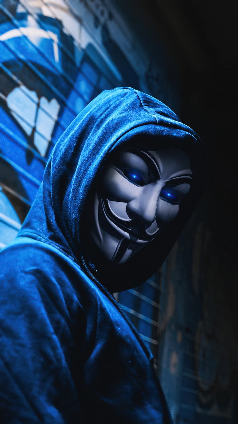 Anonymous figure in a blue hoodie wearing a Guy Fawkes mask with glowing cyan eyes against graffiti.