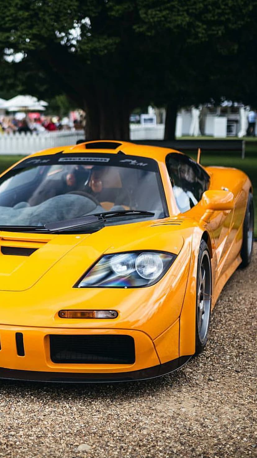 Yellow McLaren F1 supercar parked on a gravel driveway with lush green trees and white tents in the background.