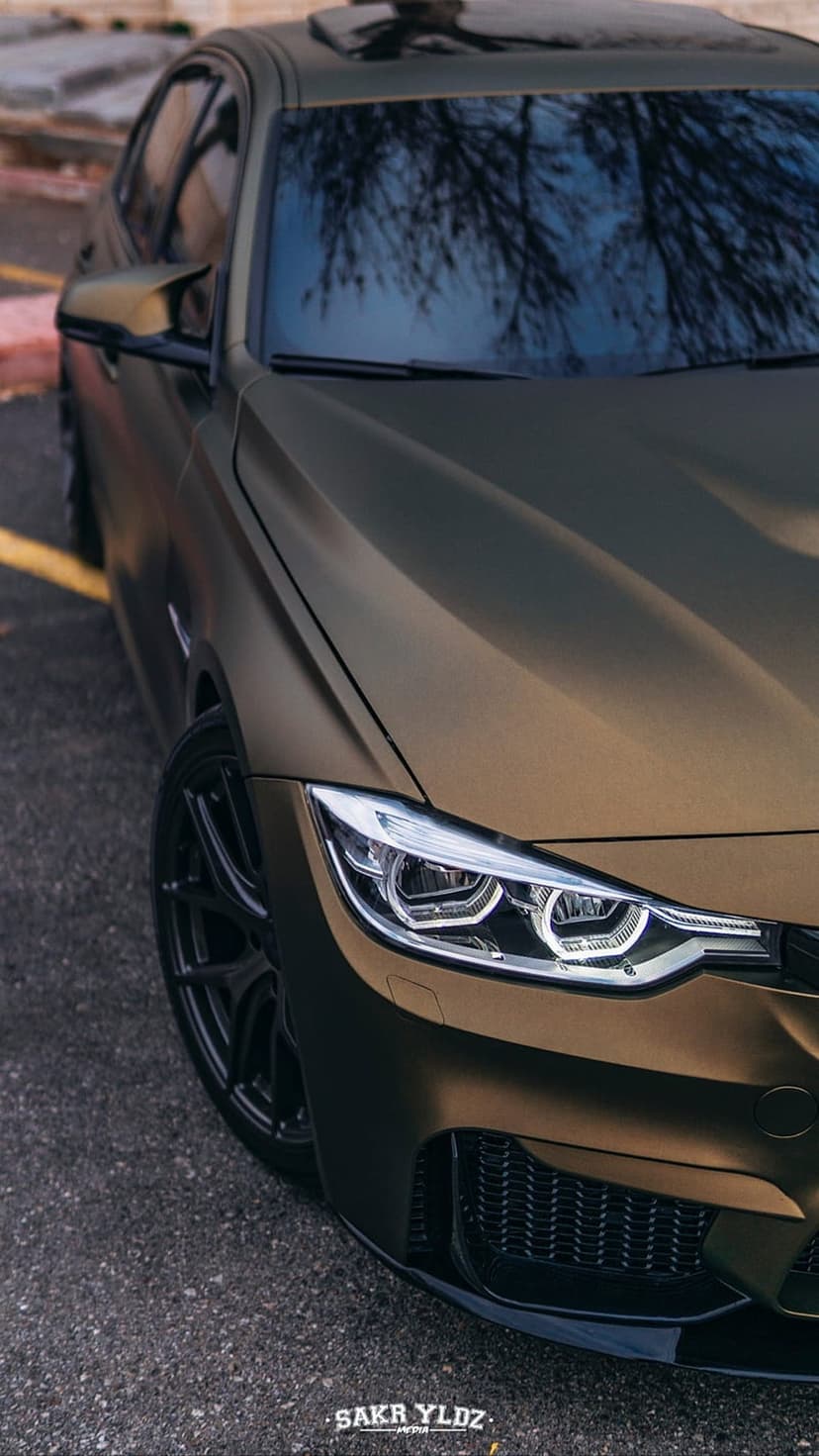 Matte bronze BMW sedan shows sharp LED headlights and black multi-spoke wheels with winter tree reflections.