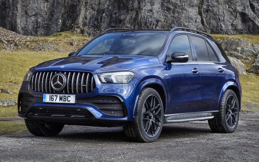 Blue Mercedes-AMG SUV parked on gravel with a rocky mountain cliff and grassy hill in the background.